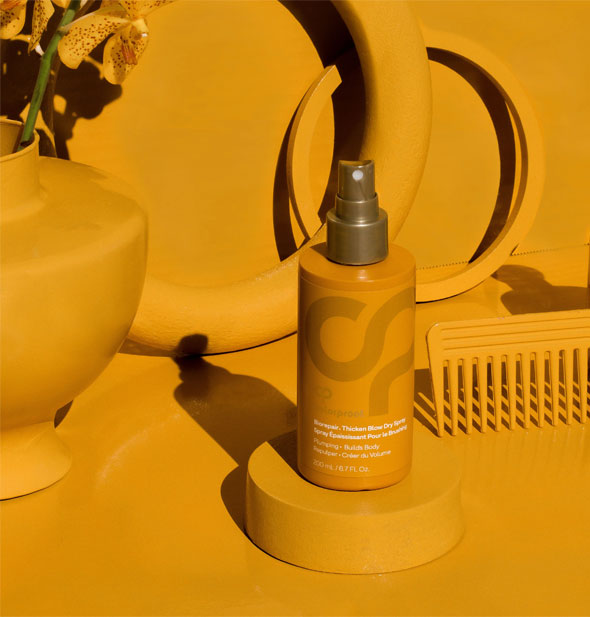 Bottle of ColorProof Biorepair Thicken Blow Dry Spray is staged with yellow flowers, hair comb, and geometric objects on a yellow background