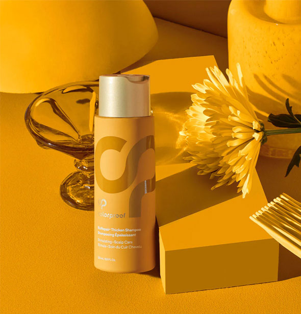 Yellow and gold bottle of ColorProof Biorepair Thicken Shampoo is staged with yellow glass, flower, hair comb, and geometric shapes 