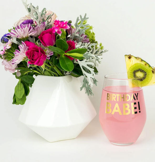 Birthday Babe! wine glass filled with a pink beverage and garnished with kiwi and pineapple slices sits next to a vase of colorful flowers