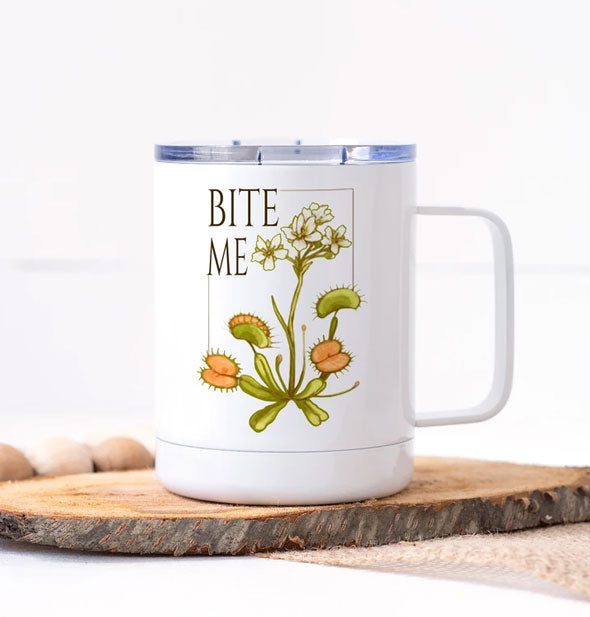 White mug with clear plastic lid and "Bite Me" artwork feautring white flowers and Venus flytraps sits on a wooden slab