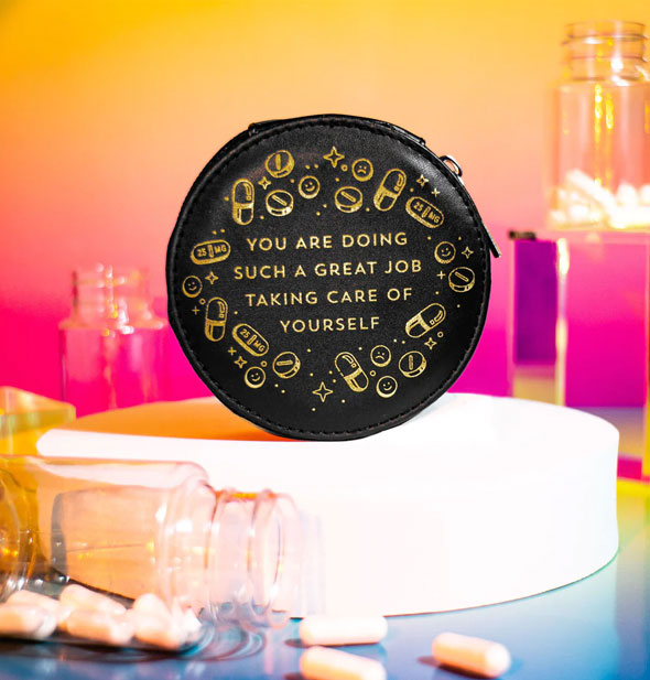 Round black pill case on a white platform surrounded by pill bottles and capsules says, "You are doing such a great job taking care of yourself" in metallic gold foil stamping surrounded by pill and star graphics