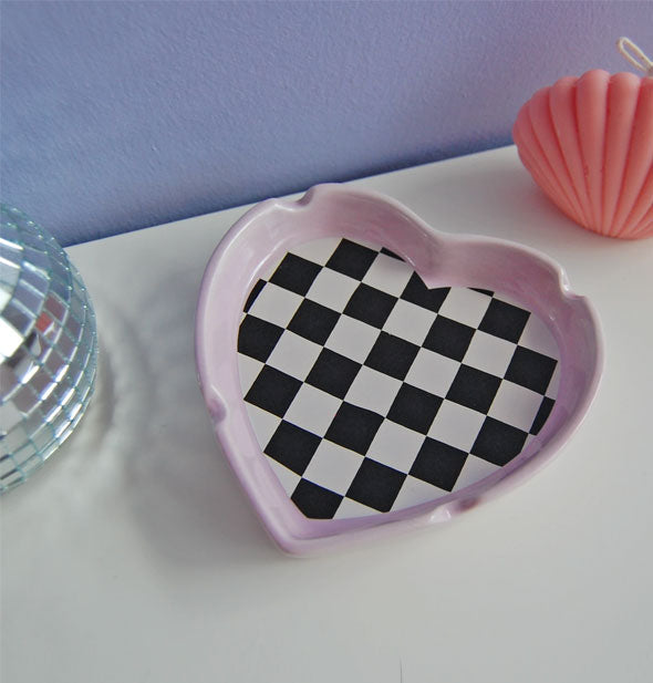 Heart-shaped ashtray with black and white checkerboard print bottom and light lavender edge sits on a tabletop with mini disco ball and coral seashell figurine