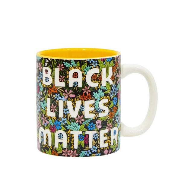 Coffee mug with white handle, yellow interior, and all-over colorful floral design says, "Black Lives Matter" in white lettering