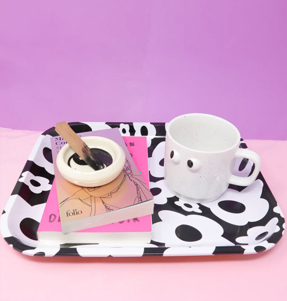 Books, a mug with googly eyes, and a partially burned palo santo stick in white yin yang dish rest on a black and white flower print tray against a pink and purple backdrop