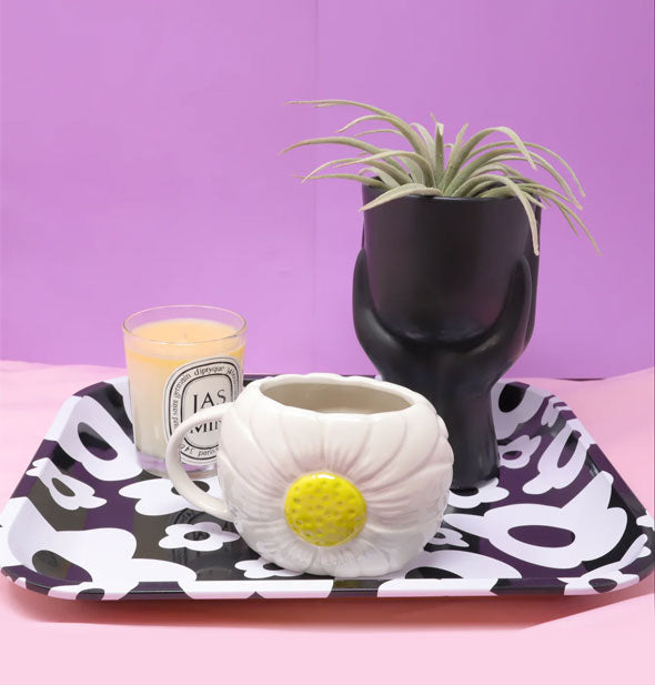 Yellow and white daisy mug, candle, and black vase holding an air plant rest on a black and white flower print tray against a pink and purple backdrop