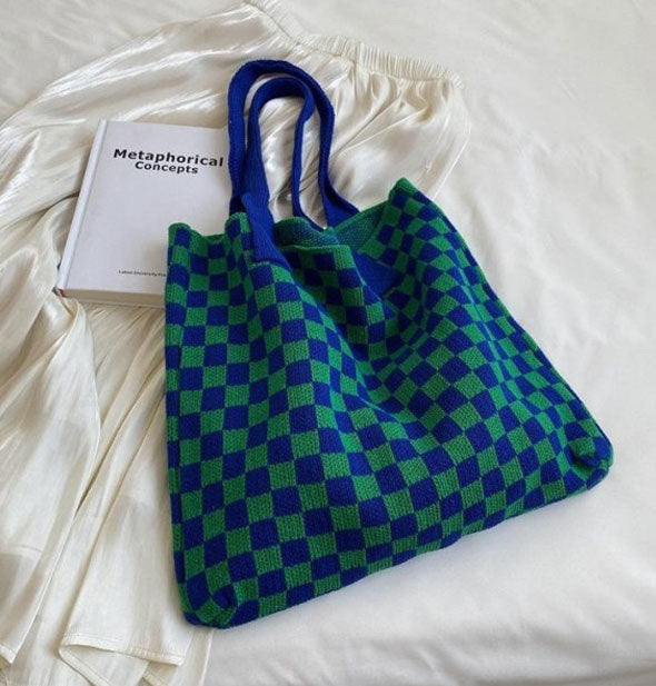 Knit blue and green checkered tote bag with blue straps rests on a white bedspread with a white satin skirt and Metaphorical Concepts book