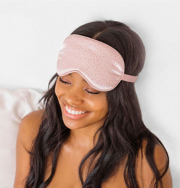 Smiling model wears a pink satin polkadot sleep mask pushed up above eyes