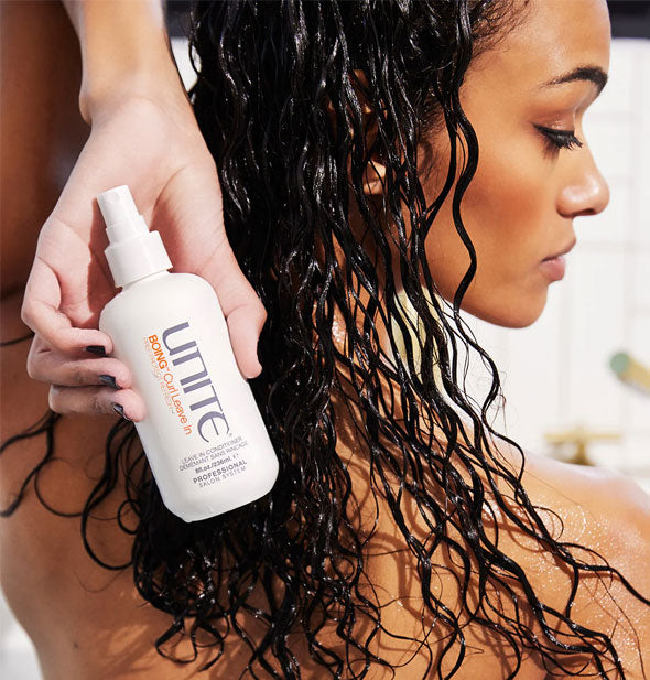 Model with well-hydrated curls holds a bottle of Unite BOING Curl Leave-In behind head