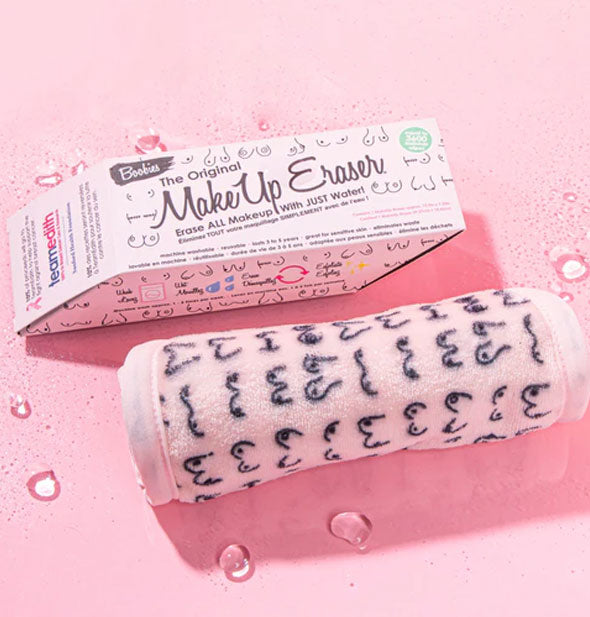 Rolled up Boobies Original MakeUp Eraser cloth with box on a pink surface sprinkled with water droplets