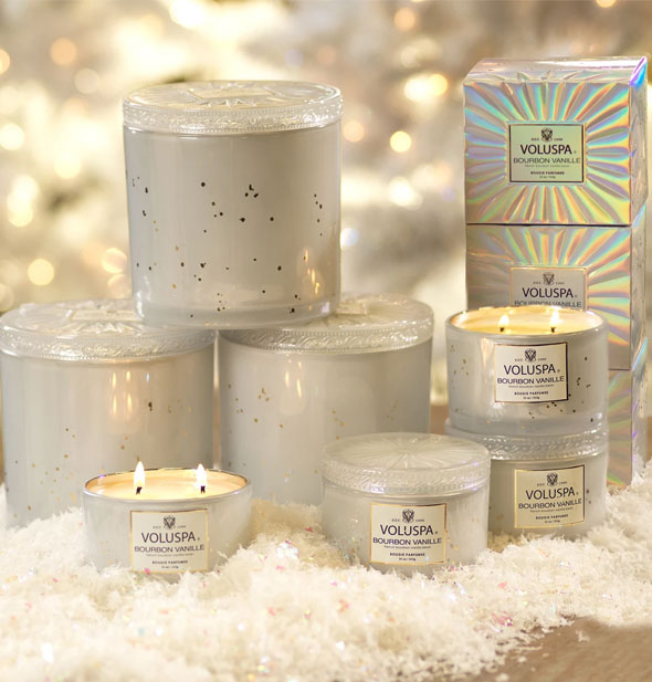Grouping of Voluspa Bourbon Vanille candles on fluffy white material, all in white speckled glass jars, most with lids, some lit, and holographic packaging shown