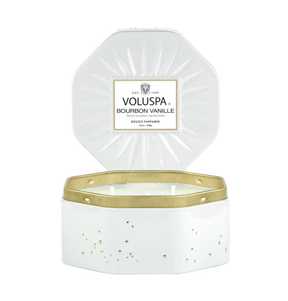 Octagonal white Voluspa tin candle with gold rim and a speckled texture with its embossed lid propped behind