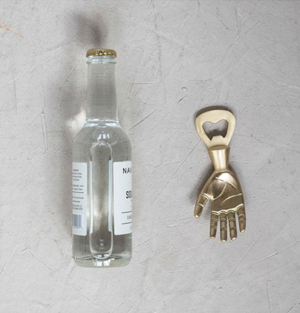 Shiny brass hand-shaped bottle opener sits next to an unopened bottle