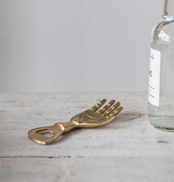Brass hand-shaped bottle opener sits on a wooden surface next to a clear glass bottle