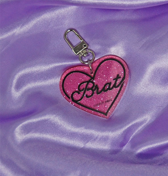 Heart-shaped pink glitter keychain says "Brat" in black script in a black heart-shaped border; silver clasp is attached