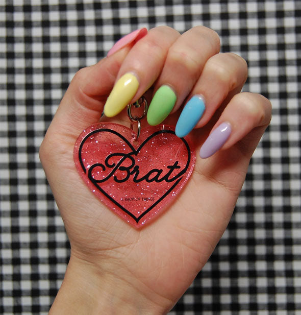 Model's hand with colorful manicure holds a heart-shaped Brat keychain against a checkered background