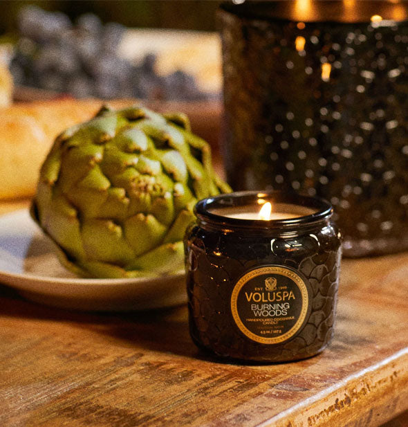 Lit Voluspa Burning Woods glass jar candle on wooden tabletop is staged with an artichoke and a larger glass vessel in the background