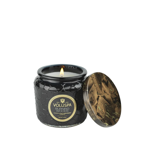 A lit Voluspa Burning Woods candle in patterned black glass jar with decorative tin lid alongside