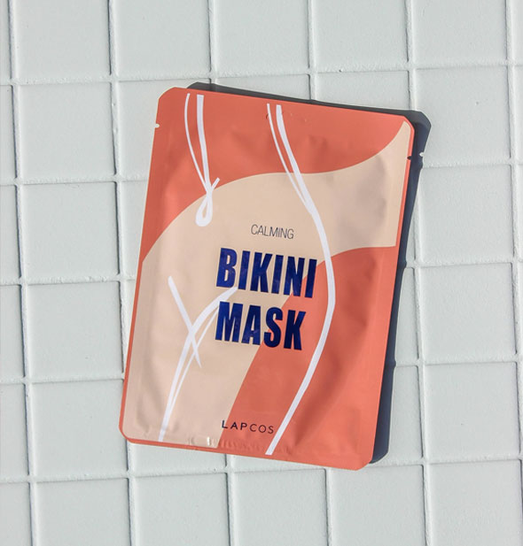 Peach and pink foil Calming Bikini Mask packet by Lapcos rests on white tile