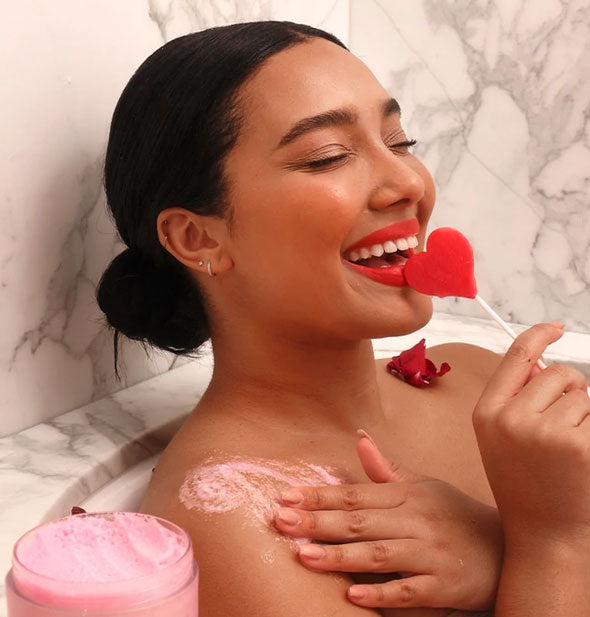 Smiling model with heart-shaped lollipop sits in a bathtub applying pink body scrub to shoulder