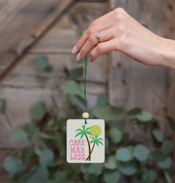 Model's hand holds a Care Way Less car air freshener by its string against a botanical and wood backdrop