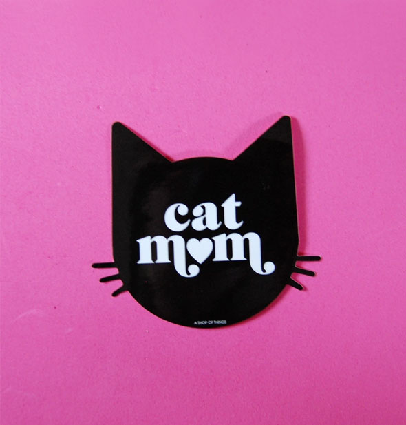 Black sticker in the shape of a cat's head with whiskers says, "Cat Mom" in white lettering with a heart in place of the "O" in "mom"