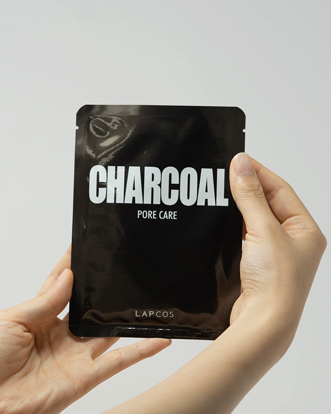 Model's hands unwrap a Charcoal Pore Care sheet mask packet and unfold the black mask inside