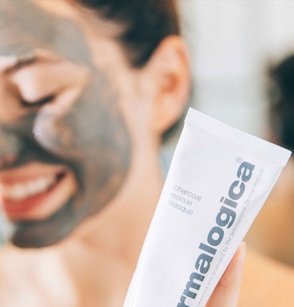 Model with Dermalogica Charcoal Rescue Masque applied to face holds a bottle of the product forward