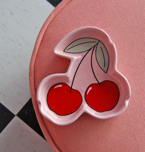 Pink ashtray shaped around a red cherries and leaves design rests on a plush pink stool on a black and white checkered tile floor