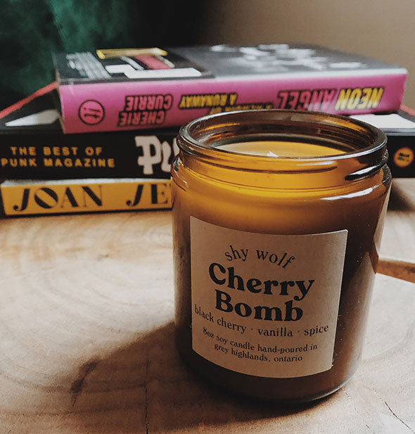 Lit Cherry Bomb candle in amber glass jar on wooden surface with a stack of books behind