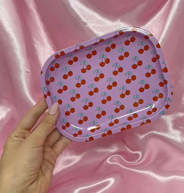 Model's hand holds a pink rectangular tray with all-over cherries pattern in front of a pink satin cloth