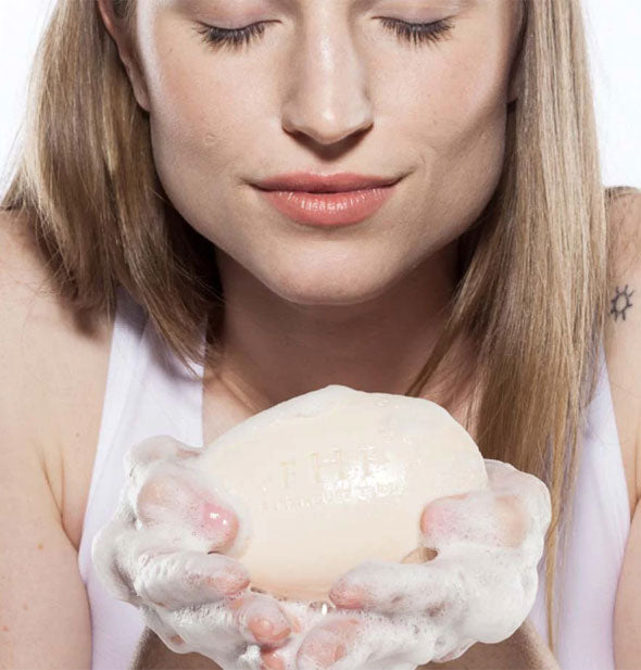 Model holds a highly lathered bar of soap in hands and leans in for a smell