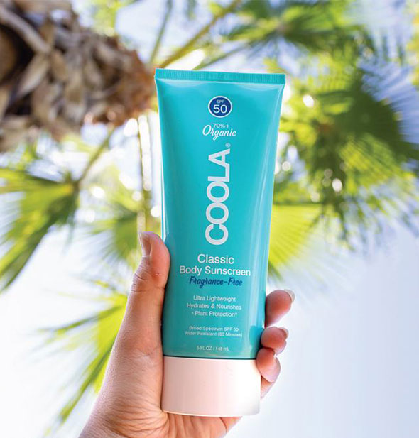 A model's hand holds a bottle of COOLA Classic Body Sunscreen in front of palm fronds and blue sky