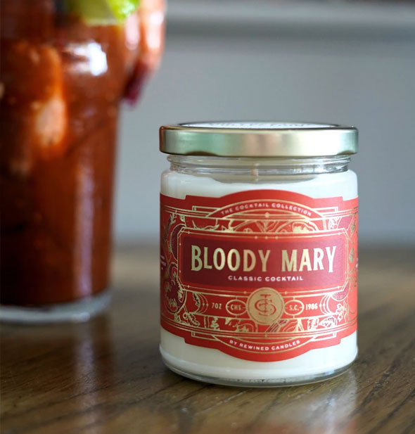 Glass jar Bloody Mary Candle with red and gold label sits on a wooden tabletop with a Bloody Mary cocktail glass in the background