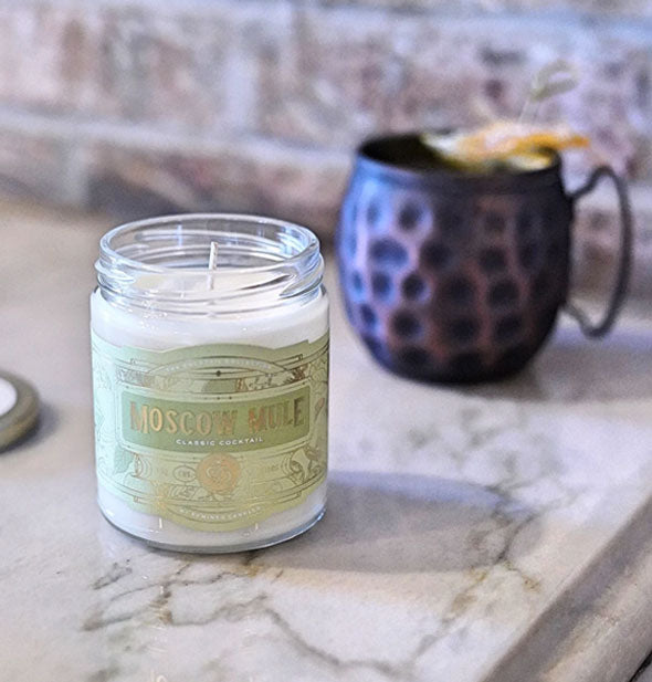 Glass jar Moscow Mule candle with green and gold label on a marble surface with blue mug in the background