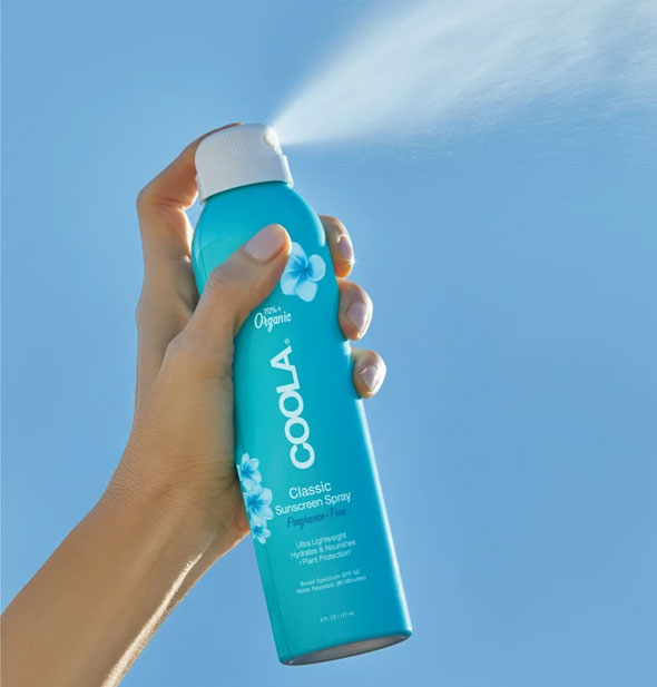 A model's hand holds and sprays from a can of COOLA Classic Sunscreen Spray in front of blue sky