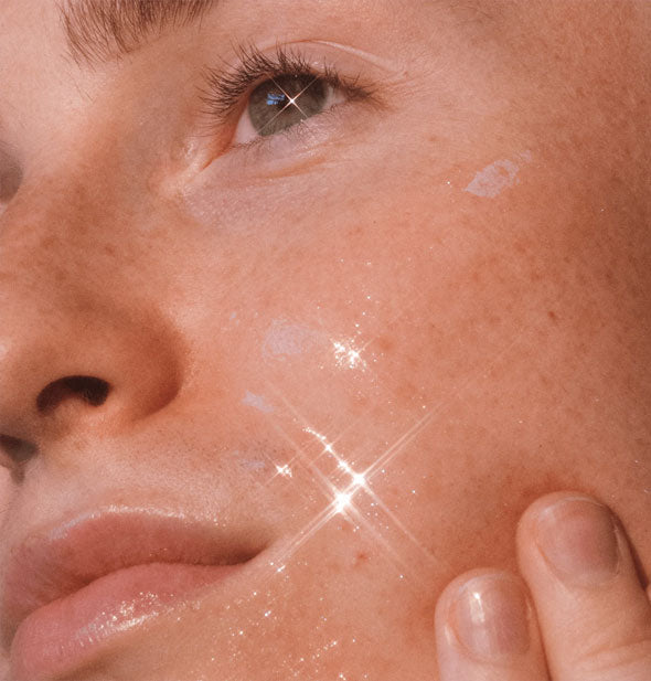 Model with dewy-looking skin that catches the light lightly touches cheek with two fingertips