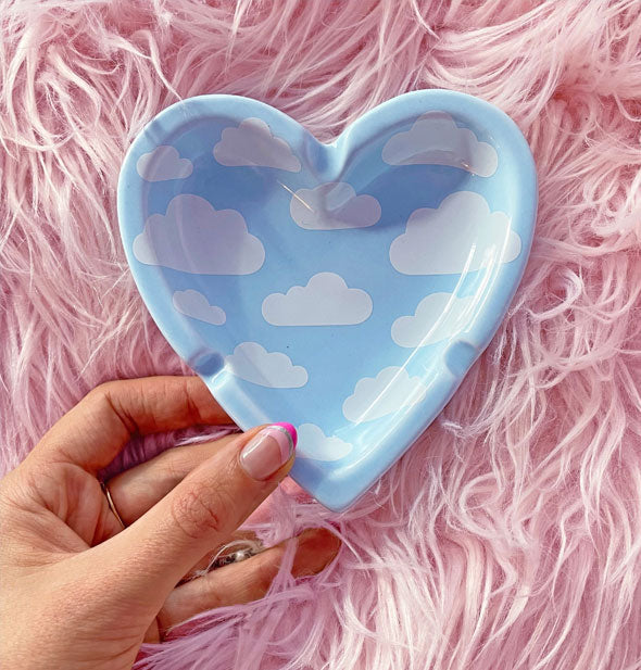 Model's hand holds a heart-shaped ashtray with all-over blue sky and puffy white clouds print in front of a pink fur backdrop