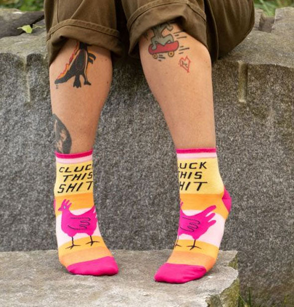 Model sitting on a large rock wears a pair of colorful Cluck This Shit socks