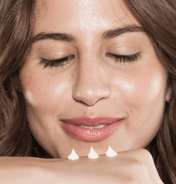 Smiling model leans in to three small dollops of cream applied to back of hand