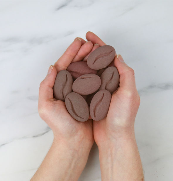 Models hands hold several coffee bean-shaped bath bombs on a marble countertop