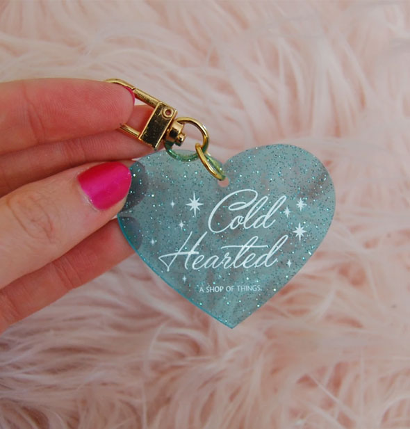 Model's hand holds a Cold Hearted blue glitter heart-shaped keychain with gold clasp