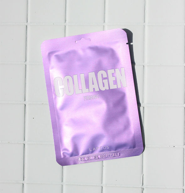 Shiny purple LAPCOS Collagen Firming sheet mask packet rests on a white tile surface