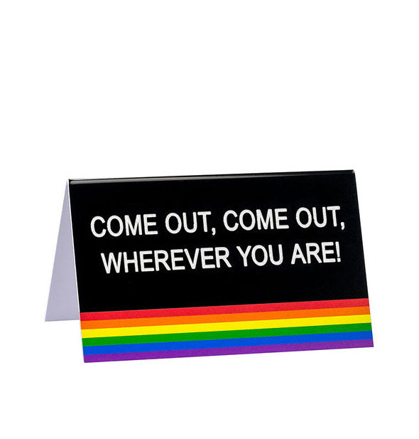 Rectangular black desk sign with rainbow stripes at bottom says, "Come out, come out, wherever you are!" in white lettering