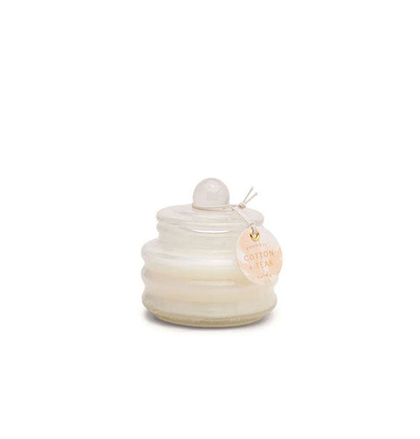 Small white ribbed glass jar candle with lid and tag noting Cotton + Teak scent