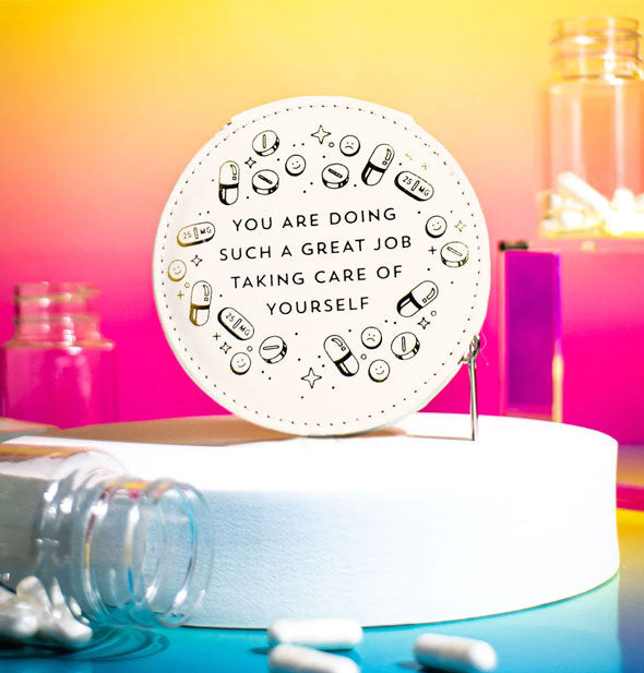 Round white pill case on a white platform surrounded by pill bottles and capsules says, "You are doing such a great job taking care of yourself" in metallic black foil stamping surrounded by pill and star graphics