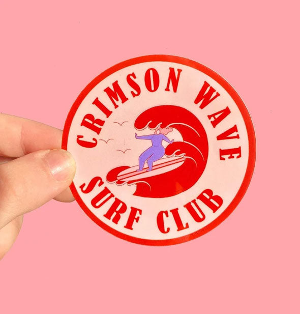 Model's hand holds a round Crimson Wave Surf Club sticker