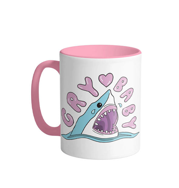 White mug with pink handle and interior features an illustration of a crying shark in water under the words, "Cry Baby" in pink bubble lettering with a matching heart graphic