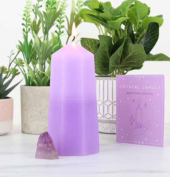 A geometrically-shaped purple candle is staged with an amethyst crystal, purple Crystal Candle booklet, and houseplants in the background on a white marble surface