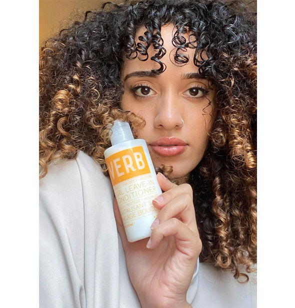 Model with very curly hair holds a bottle of Verb Curl Leave-In Conditioner