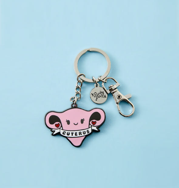 Pink enamel keychain charm with silver hardware attached resembles a smiling cartoon uterus with hearts on the ends of its fallopian tubes, wrapped in a white banner that says, "Cuterus"